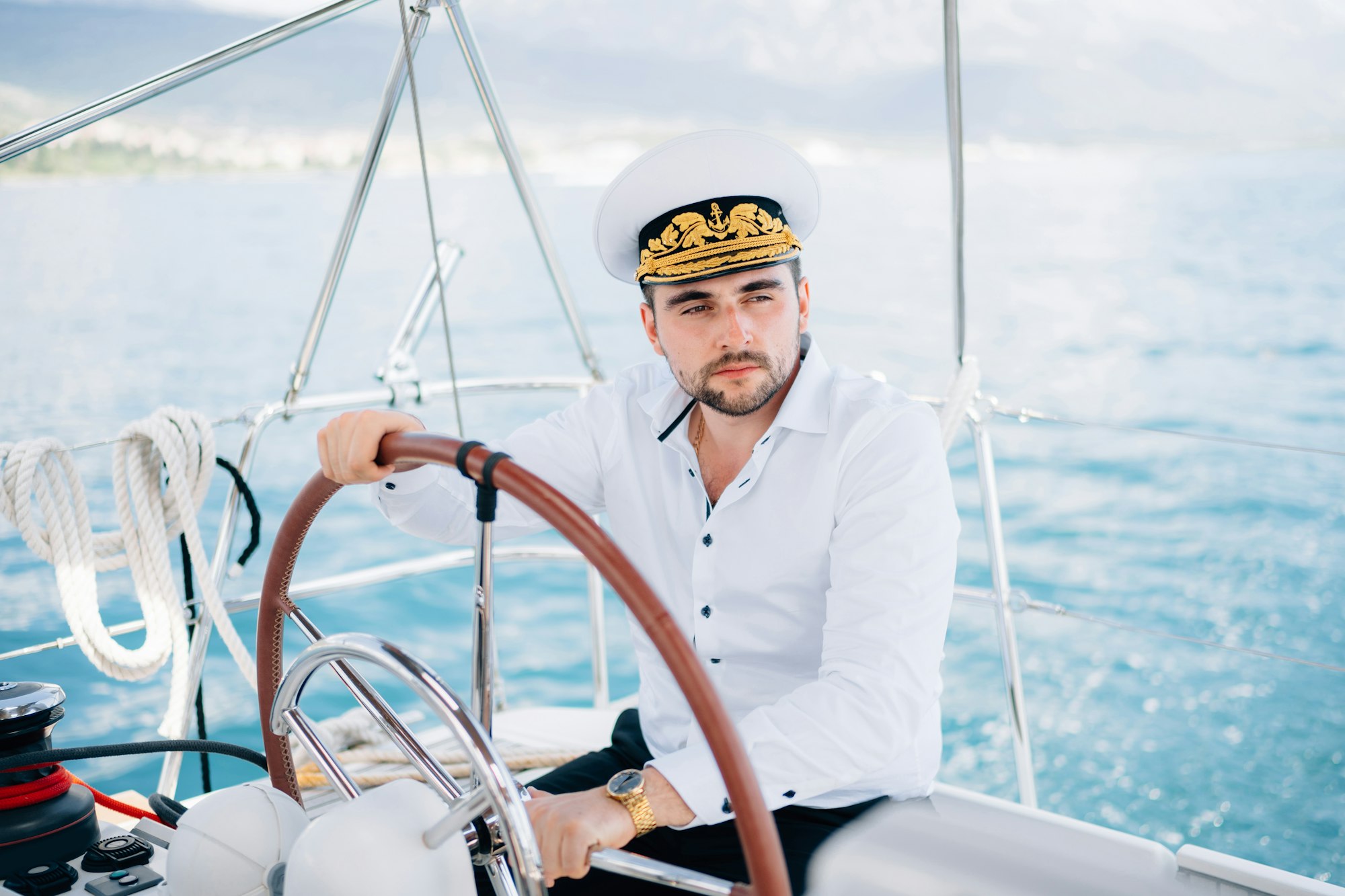 A man in a captain's cap sits at the helm on a yacht sailing in the middle of the sea